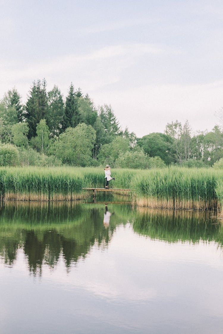 Couple Standing On Wooden Dock Standing On Green Grass Field Near Lake