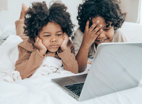 Free Uninterested ethnic boy and positive brother in pajamas lying on stomachs on bed while using laptop browsing Internet Stock Photo