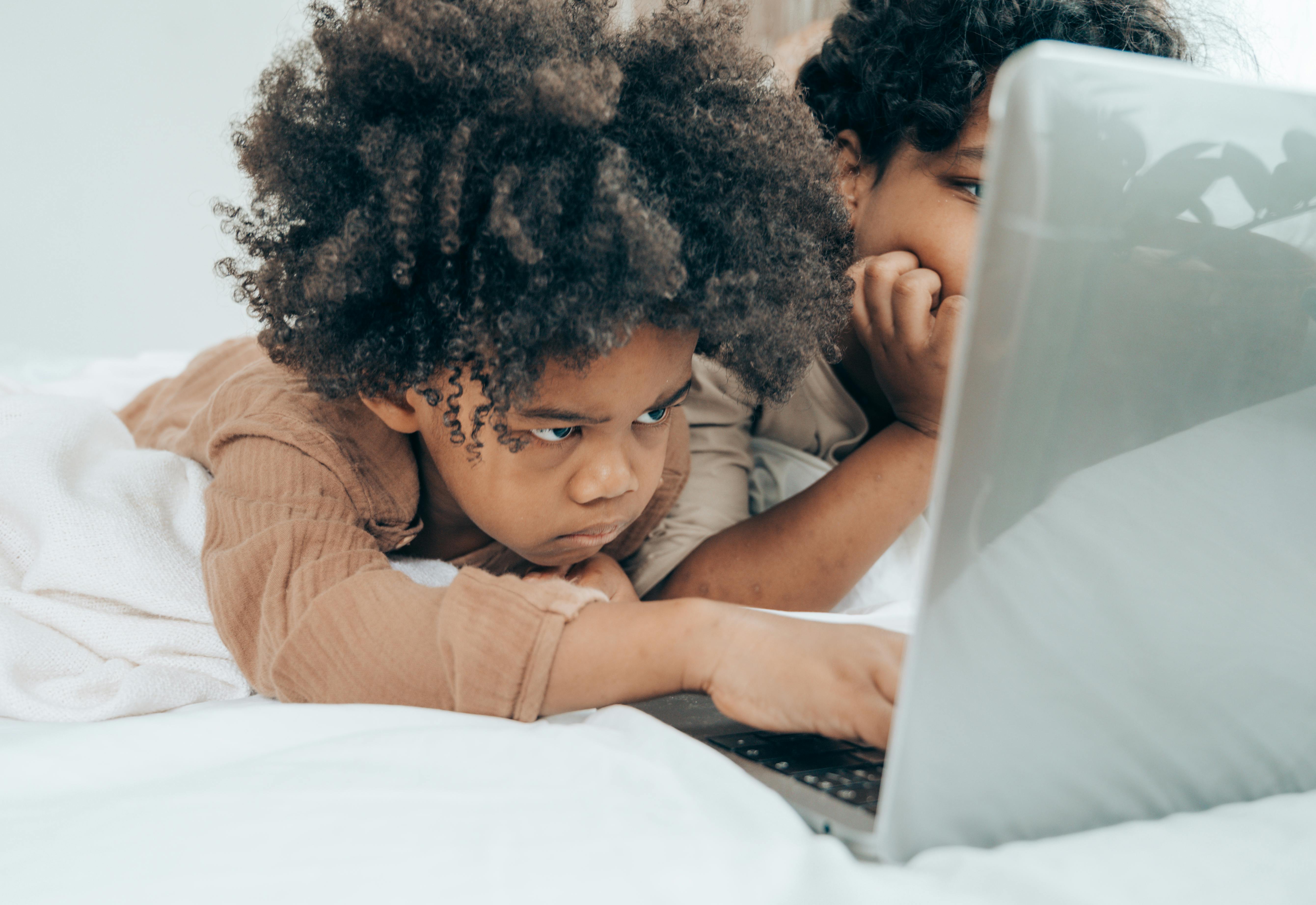 focused black boy using laptop with brother on bed