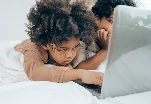 Concentrated African American kid in pajama using laptop with brother while lying on bellies on bed in light room