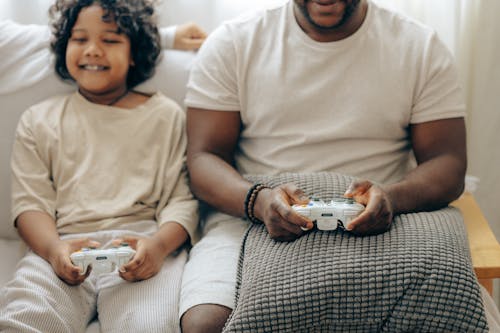 Crop smiling black father and son in sleepwear playing with joysticks on game console together while sitting on sofa in light living room