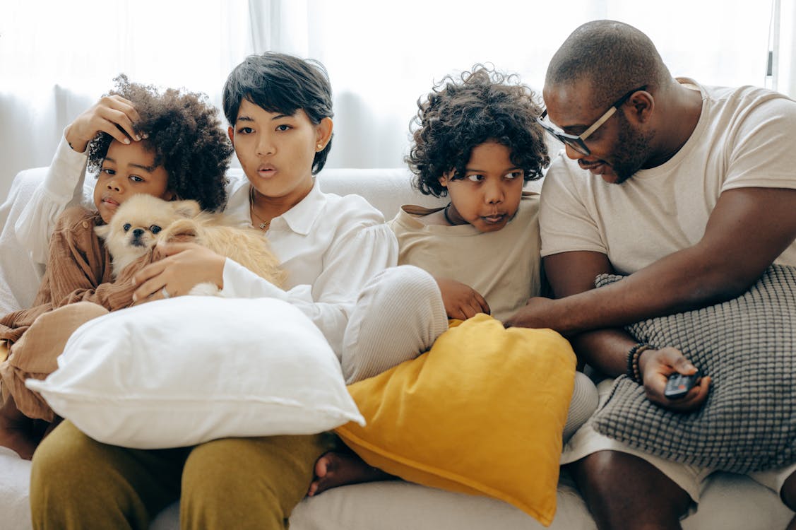Free Multiracial family resting on comfortable sofa together with little dog while mother and little son watching TV with interest and father and elderly son talking to each other Stock Photo