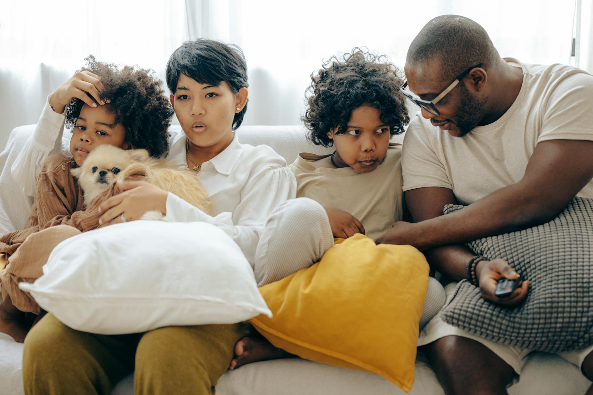 Multiracial family resting on comfortable sofa together with little dog while mother and little son watching TV with interest and father and elderly son talking to each other