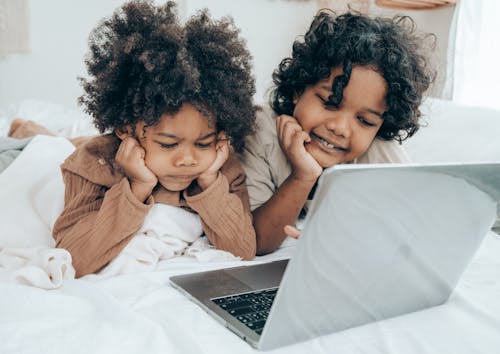 Free Disappointed and smiley black brothers in pajamas lying on bed in daytime while using laptop surfing Internet together Stock Photo