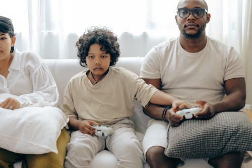 Free Concentrated child showing usage of gamepad to father while playing on game console and resting on sofa together with family Stock Photo