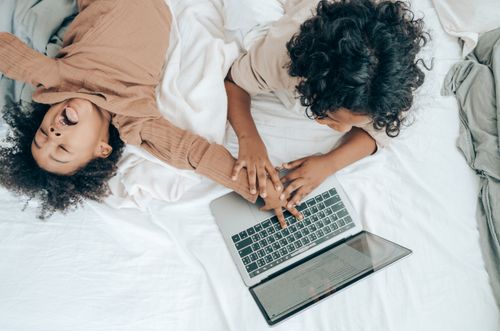 Free From above of black kid lying on bed and fooling around brother typing on laptop keyboard while brother reading document Stock Photo