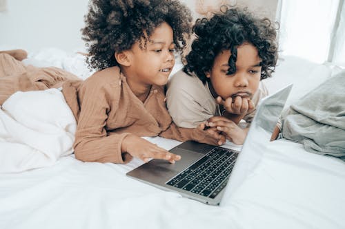Side view of happy and sad ethnic little brothers in sleepwear lying on bellies on bed in light room together while surfing Internet on laptop