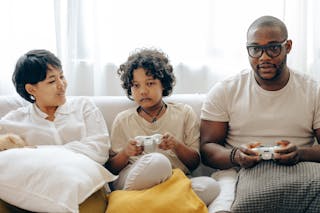 Focused ethnic child and dad in sleepwear using gamepads for playing video games on console via TV while sitting on comfortable couch together with mom