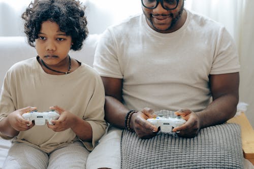 Free Crop ethnic male parent and son sitting near each other on sofa while using playstation and joysticks playing video games Stock Photo