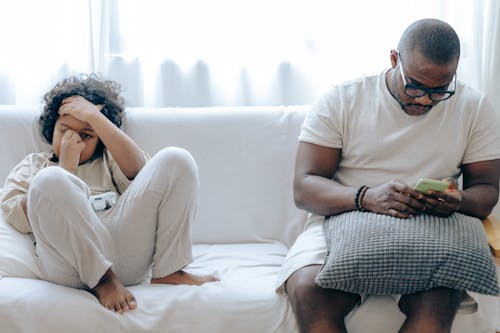 Free Concentrated African American man in nightwear using smartphone while sitting on cozy sofa with son resting with joystick in light room Stock Photo