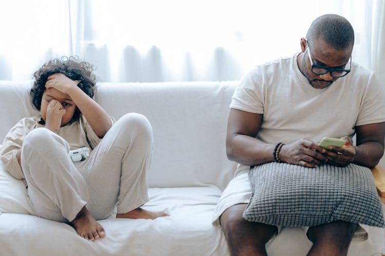 Black Man Using Smartphone On Sofa With Son Sitting Together