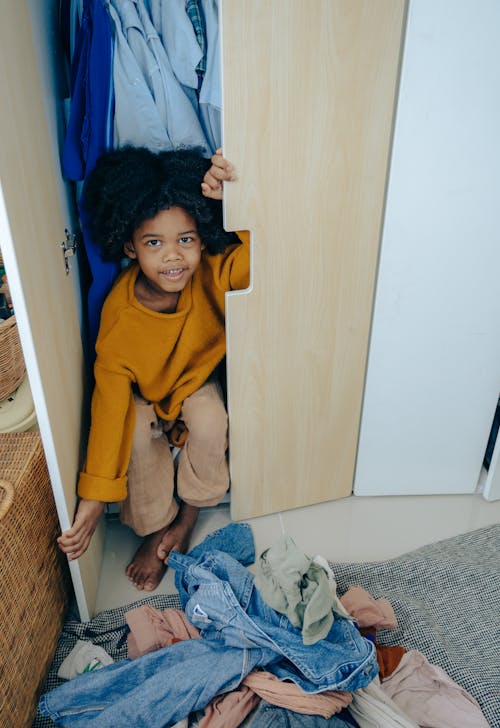 Playful little African American kid sitting in wardrobe in bedroom