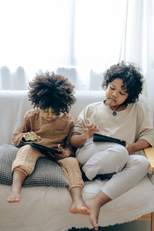Free Adorable little ethnic sisters sitting together on comfortable sofa and playing with tablets while spending time at home Stock Photo