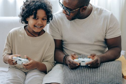 Free Cheerful crop African American child playing video game with joystick together with father sitting on sofa at home Stock Photo