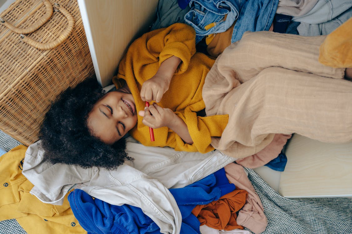 Free Top view of funny cute little African American girl lying on pile of scattered clothes and drawing on hand with marker Stock Photo