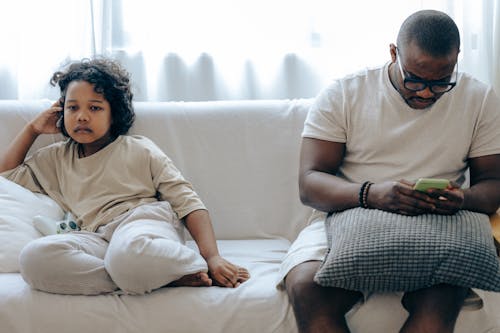 Young African American man surfing internet on mobile phone sitting on sofa together with daughter watching TV