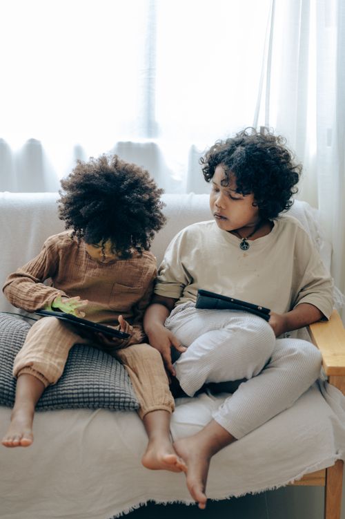 Free Full body of adorable little black siblings in pajamas chilling on sofa and playing game on tablet together in cozy living room Stock Photo