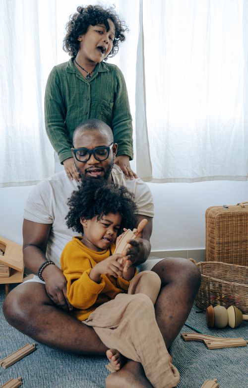 Free Positive young black man in comfy clothes and eyeglasses sitting on floor in room and playing with adorable daughters Stock Photo
