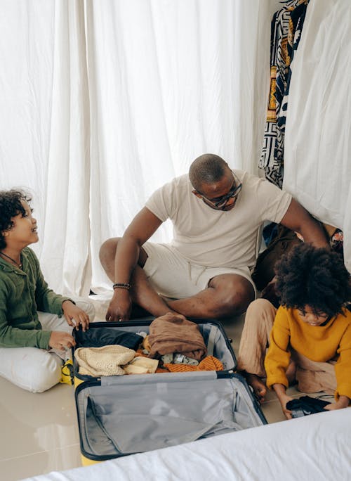 From above of young African American man sitting on floor and packing suitcase with little daughters