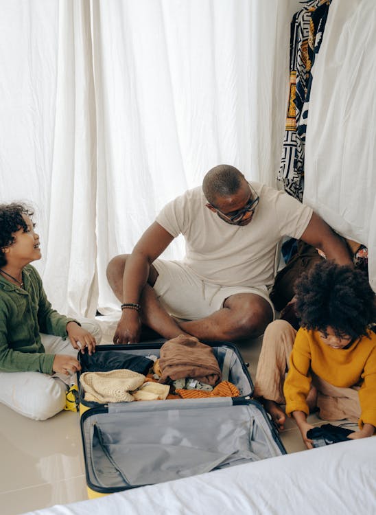 Free From above of young African American man sitting on floor and packing suitcase with little daughters Stock Photo