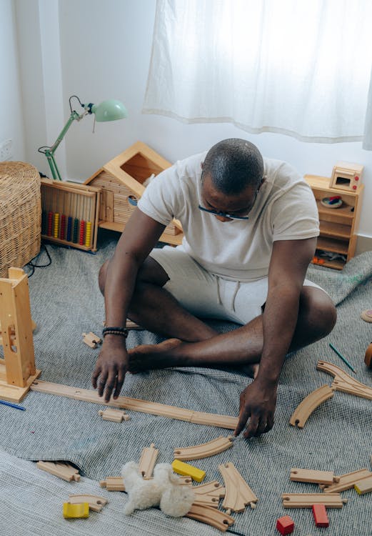 From above of African American male in glasses and casual wear sitting on floor near unfinished wooden construction