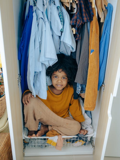 Content African American child with curly hair sitting in wardrobe among cloth and looking at camera