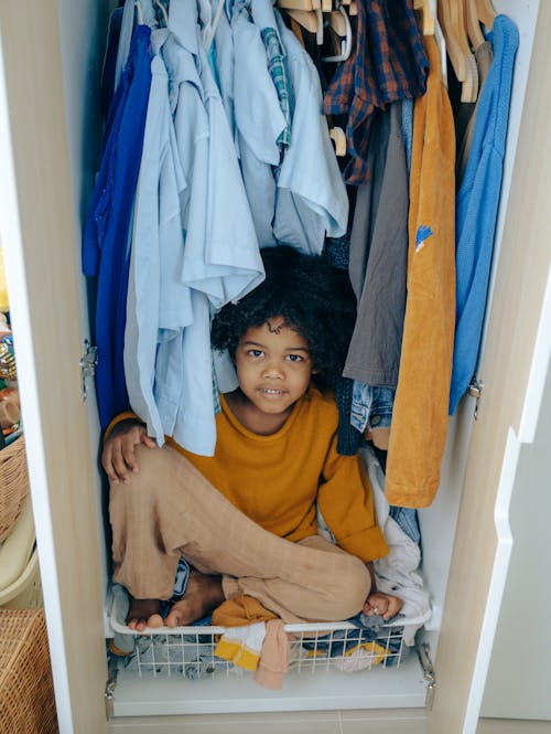 Free Content African American child with curly hair sitting in wardrobe among cloth and looking at camera Stock Photo