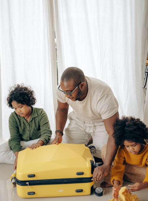 African American male in eyeglasses sitting with sad children with curly hair on floor and packing suitcase