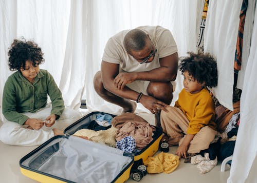 Free African American father in eyewear with sad kids with curly hair sitting near opened suitcase Stock Photo