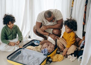 African American father in eyewear with sad kids with curly hair sitting near opened suitcase