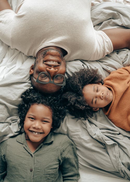 Top view of African American siblings with curly hair and father in eyeglasses lying on bed