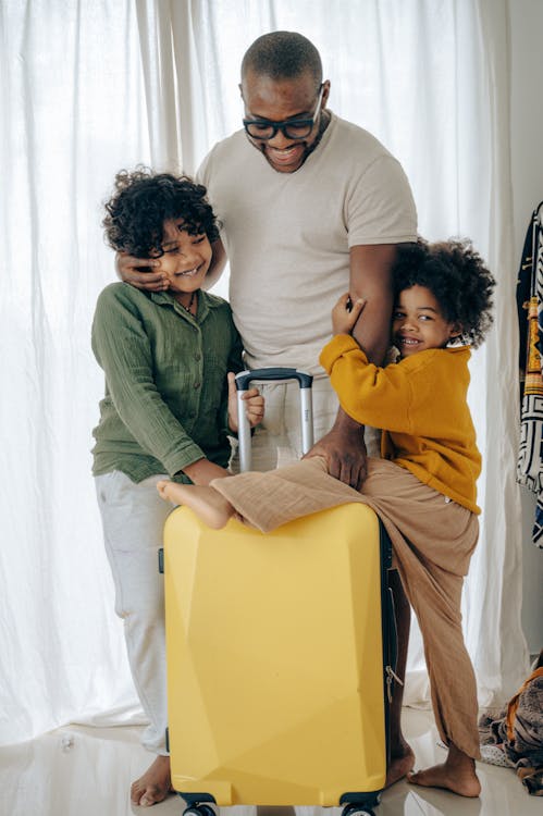 Free Positive African American man in eyewear hugging with with kids with curly hair and standing near suitcase Stock Photo