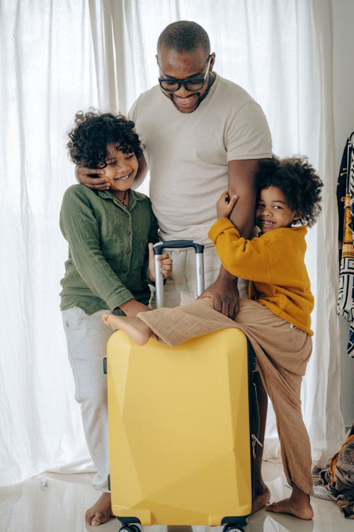 Positive African American man in eyewear hugging with with kids with curly hair and standing near suitcase