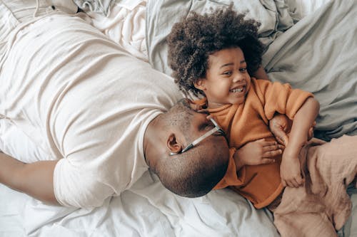 From above of cheerful African American man in eyeglasses and child with curly hair in sleepwear lying on bed