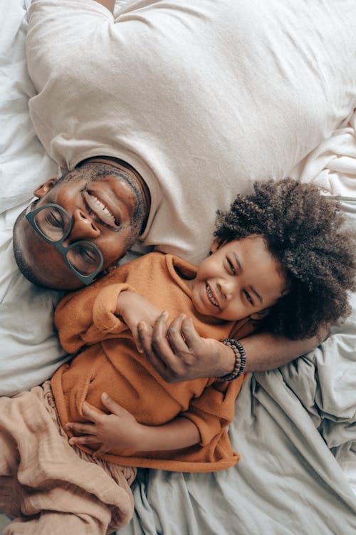 Top view of excited African American male in glasses and kid with curly hair lying on bed next to each other