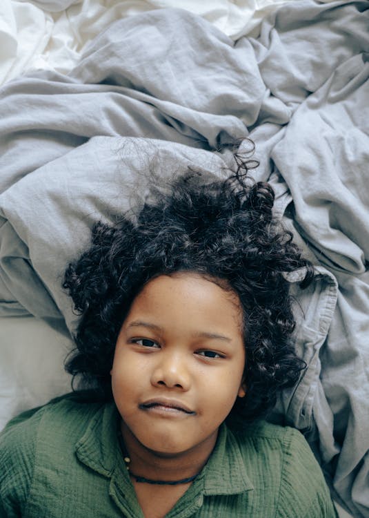 Top view of African American child with dark curly hair in casual clothes lying on rumpled bed and looking at camera while resting in comfortable apartment