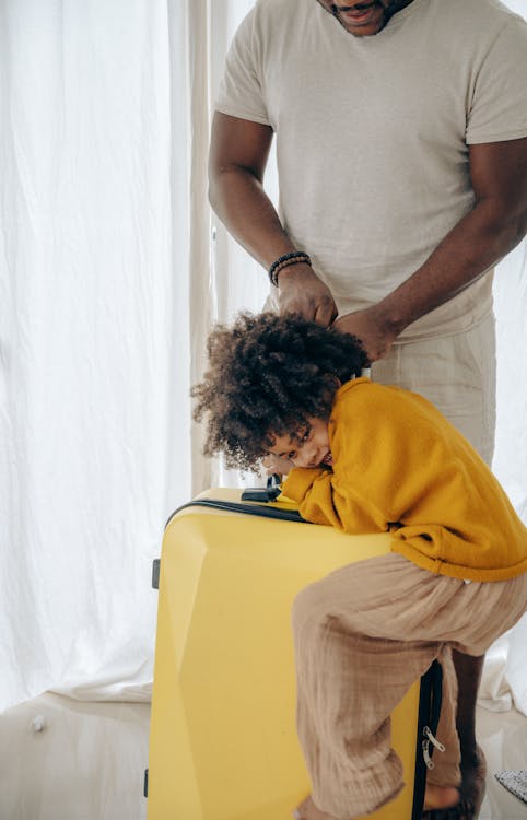 Free Unrecognizable African American cute curly child clutching large yellow suitcase while man in casual clothes going to leave from home Stock Photo