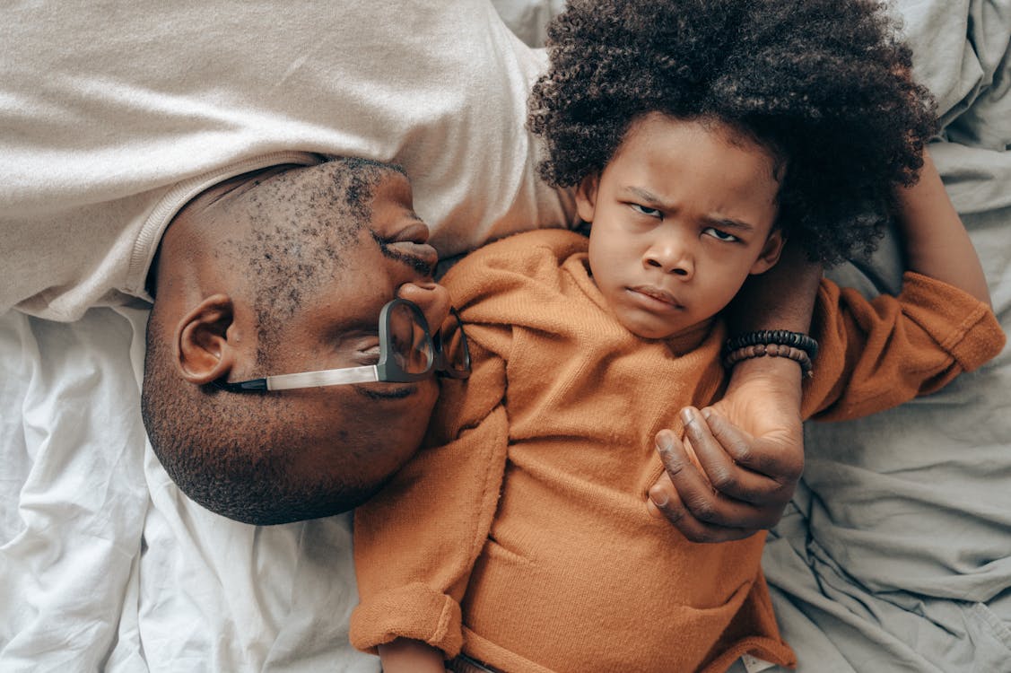 Top view of African American man in glasses lying near angry child in casual clothes while cuddling together in comfortable bedroom