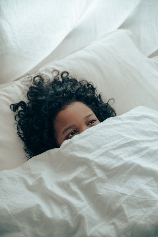 From above of unrecognizable ethnic kid with curly hair lying in white comfortable bed while covering lower part of face with  white blanket in light room