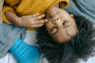 一个穿着黄色毛衣、面无表情的民族儿童躺在床上，从上方可以看到他穿着牛仔裤和蓝色袜子的腿