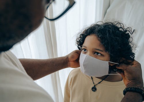 Free From above unrecognizable black man wearing protective mask on face of ethnic kid standing in room near window closed with curtain Stock Photo