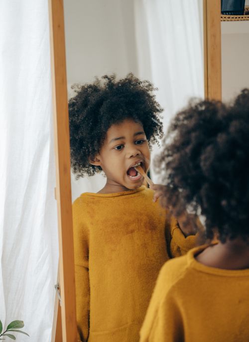 Free Ethnic child in yellow sweater using toothbrush for cleaning teeth while standing in bathroom looking at mirror Stock Photo