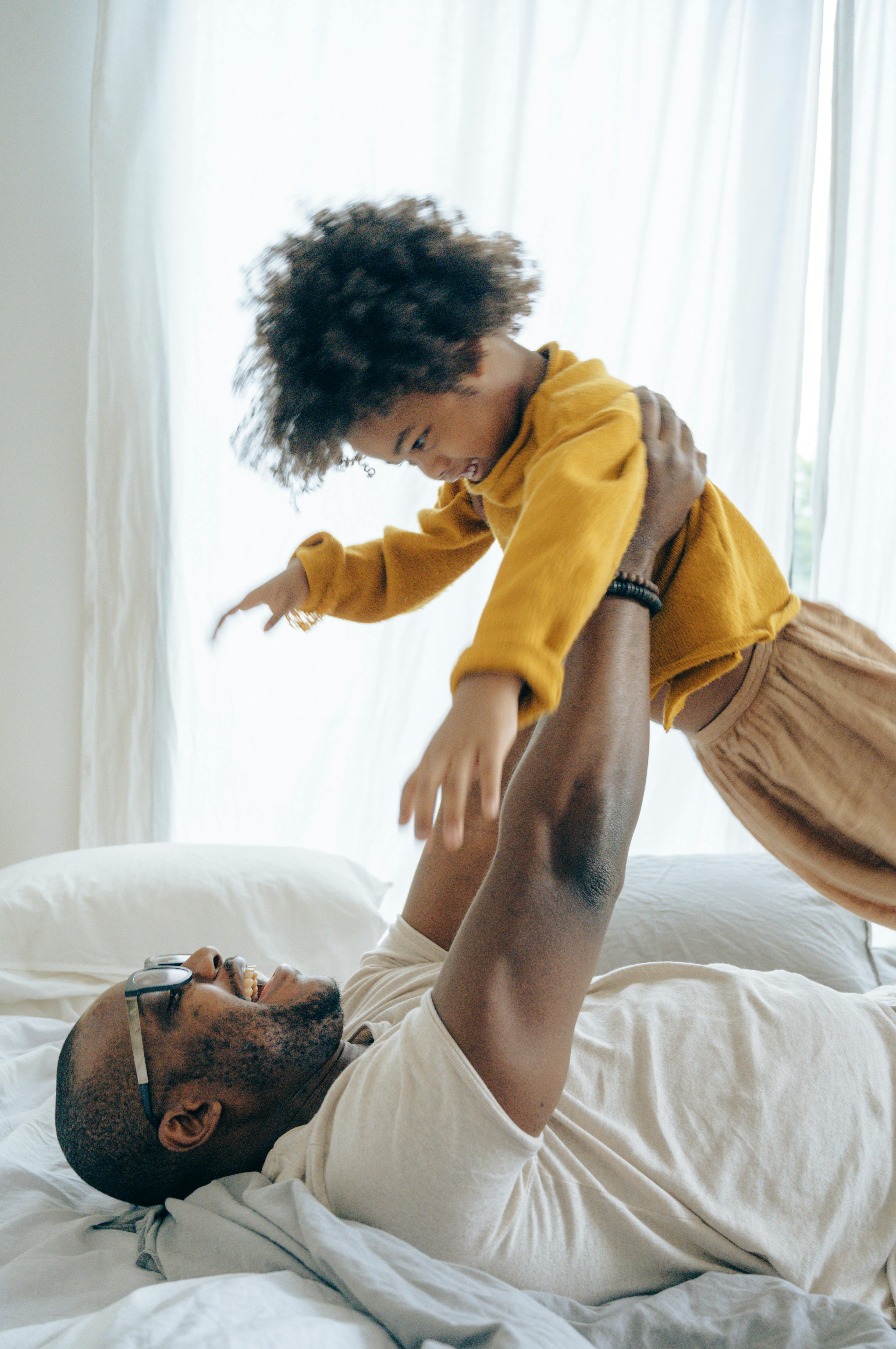 laughing black man raising kid up lying on bed