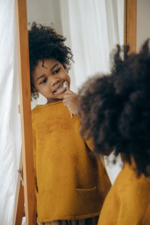 Concentrated black child in yellow sweater cleaning teeth with toothbrush looking at long mirror in bathroom