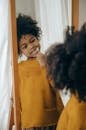 Concentrated black child in yellow sweater cleaning teeth with toothbrush looking at long mirror in bathroom