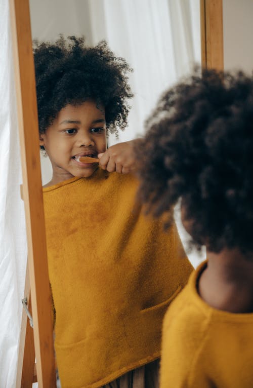 From behind of crop positive adorable black boy in orange casual wear brushing teeth against narrow mirror in wooden frame in light bathroom