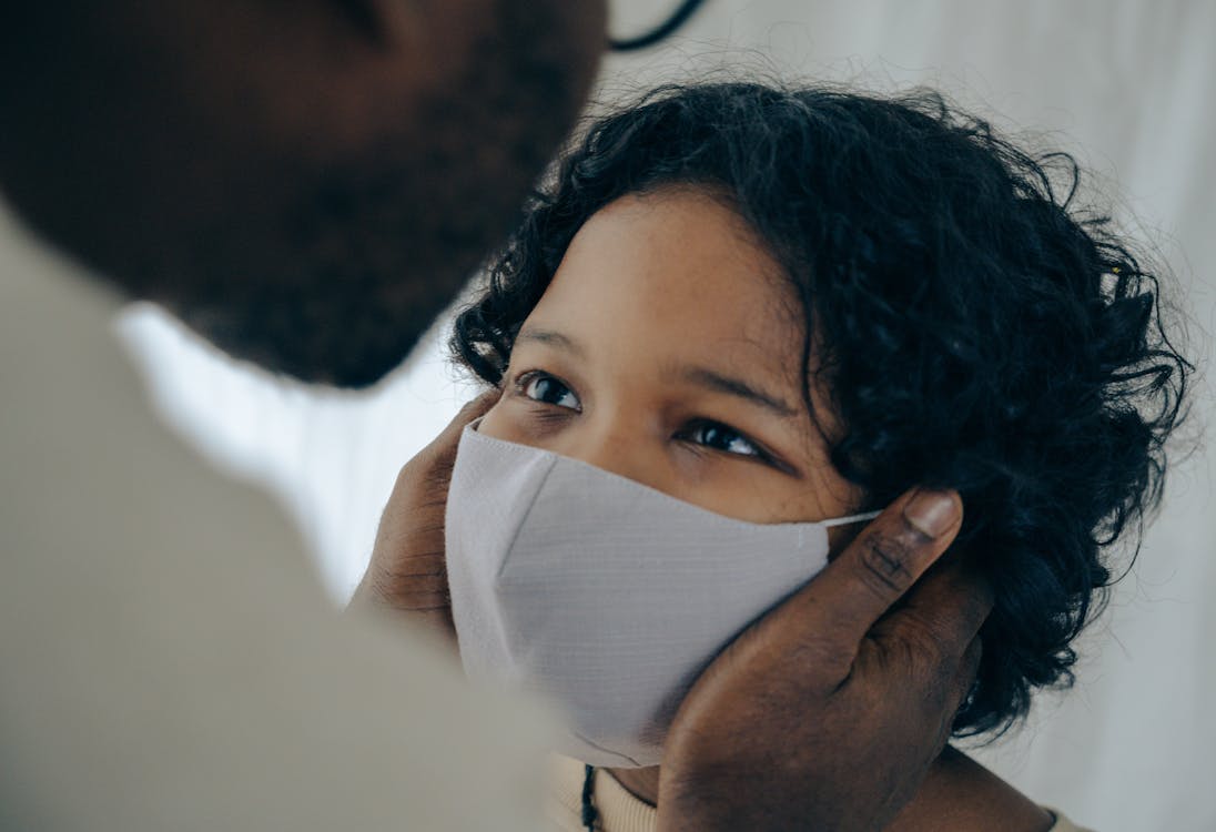 Black man in eyeglasses gently touching cheeks of cute calm little son wearing respirator during coronavirus pandemic