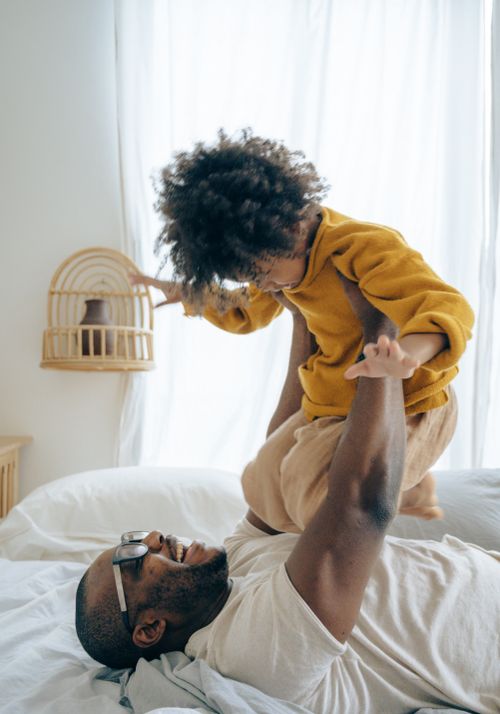 Free Side view happy black man in eyeglasses lying on comfy bed and lifting up adorable son in pajama while having fun together in cozy light bedroom in morning Stock Photo