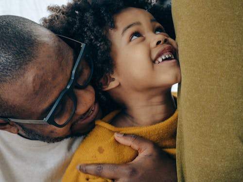 Crop cheerful African American man in casual shirt and eyeglasses hugging cute smiling son with curly hair in daytime