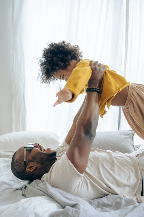 Free Side view bearded black father in white casual shirt resting on cozy bed with white sheets and lifting up happy little boy in orange pajama after awakening in light bedroom Stock Photo